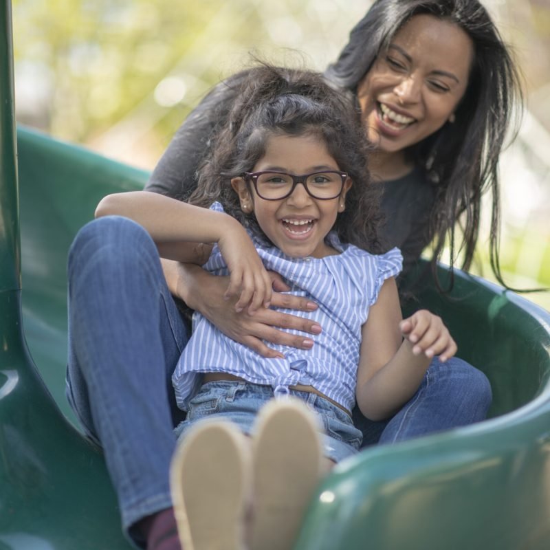 A young hispanic mother and daughter are playing together at the park. They are sliding down a slide and laughing with excitement. They are wearing casual clothes and the young child is wearing reading glasses.